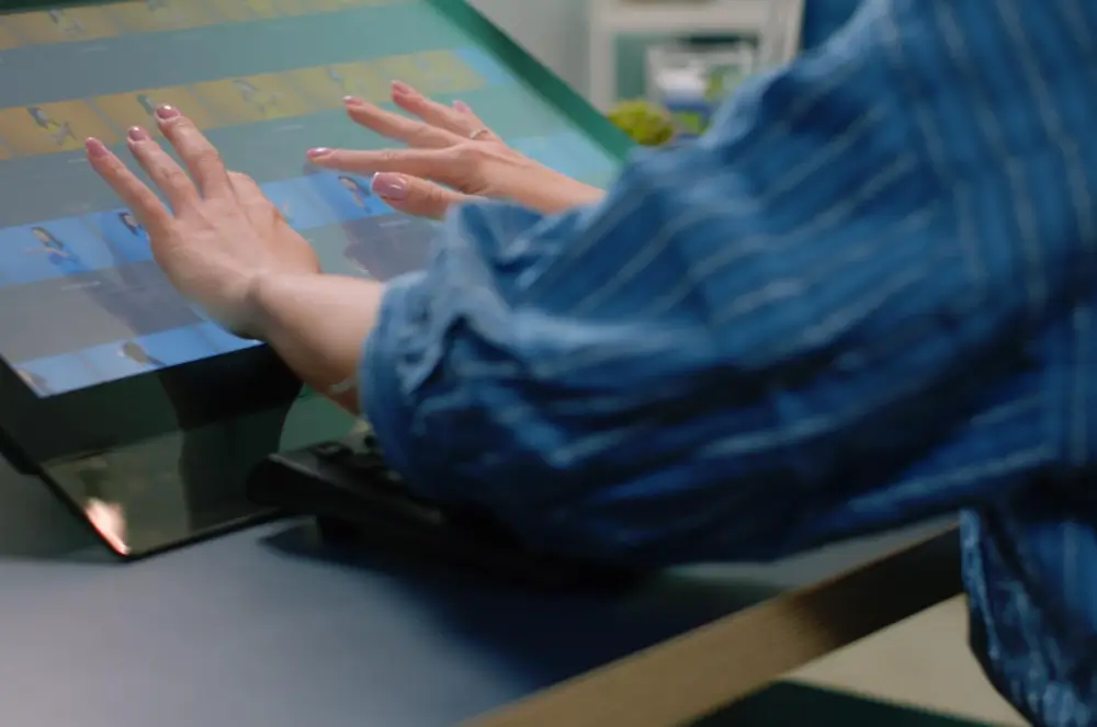 A person in a blue shirt interacts with a digital touch screen displaying colorful squares and lines. Their hands are extended towards the screen, suggesting engagement with the interface.